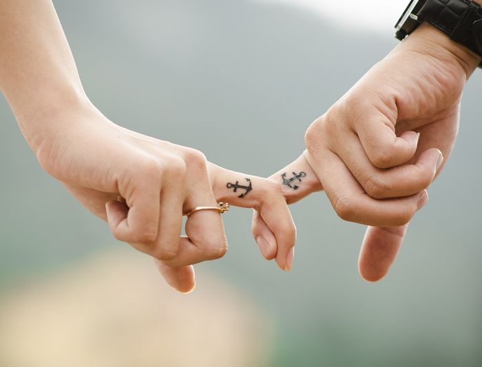 Two persons holding hands. They both have a tattoo depicting an anchor on their index finger.