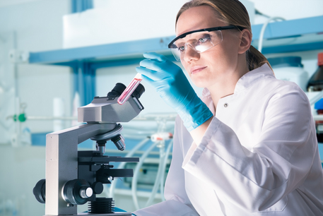 Junge Frau im weißen Kittel, mit Schutzbrille und blauen Laborhandschuhen steht im Labor und betrachtet eine rote Flüssigkeit in einem Reagenzglas, dass sie in der Hand hält. Neben ihr steht ein Lichtmikroskop, im Hintergrund steht eine Laborbank mit Absaugung. 