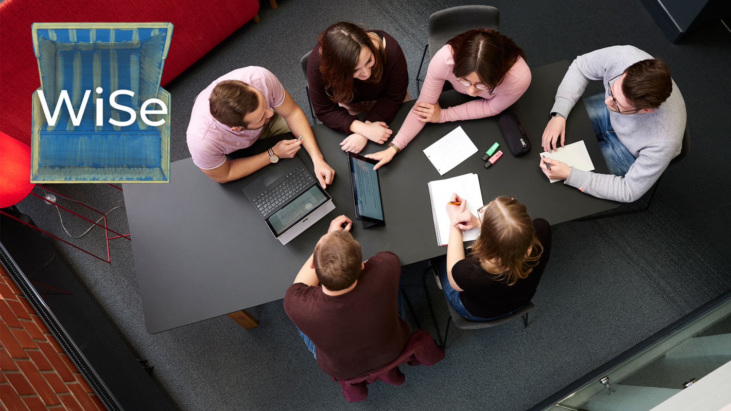 Eine Gruppe von Studierenden sitzt um einen Tische herum und unterhält sich angeregt. Die Gruppe ist von oben fotografiert.